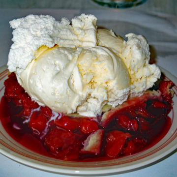 A giant slice of cherry pie occupies a full plate and is topped with a half-pint of vanilla ice cream.