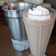 Tall soda fountain glass holds a whipped cream-topped malt, with a silver beaker in back for refills.
