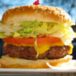 Cheeseburger is stacked in a bun with tomato and lettuce on a sunny outdoor table.