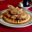 Pieces of fried chicken are piled atop a waffle, pitcher of syrup in the background