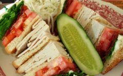 Quartered club sandwich features turkey, tomato, and lettuce, accompanied by a pickle spear and cole slaw