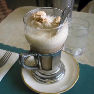 On a lunch-counter place mat, soda fountain glass with a metal handle contains a coffee ice cream soda