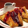 Six thick half-slices of bread, dipped in egg batter and fried, are arrayed on a plate under fruit jam, with a pitcher of syrup on the side.