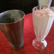 Strawberry shake in a tall soda fountain glass is accompanied by a silver beaker of extra milk shake for refills