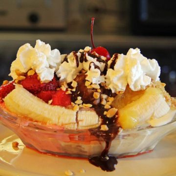 Glass soda fountain boat holds scoops strawberry, chocolate, and vanilla ice cream topped with sauce, nuts, and whipped cream, shored in by bananas sliced lengthwise