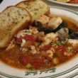 Bowl full of seafood in herbed tomato sauce, accompanied by toasted sourdough bread