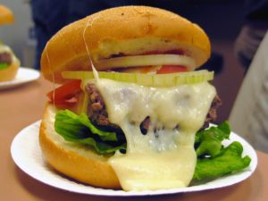 Molten cheese oozes off a tender burger in a bun with lettuce, tomato, and sliced onion at Ted's in Meridian, CT