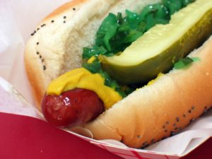 Poppy seed bun holds hot dog with mustard, relish, and pickle spear