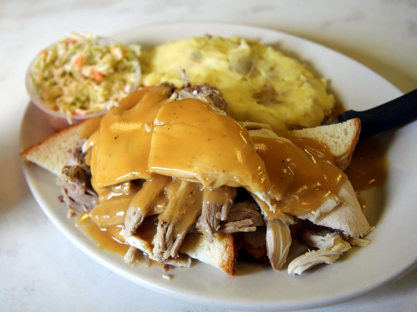 Gravy spills across a pile of just-carved turkey in an open-face fandwich, with sides of mashed potatoes and cole slaw.