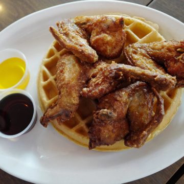 Pieces of fried chicken are arrayed atop a round Blgian waffle, sided by melted butter and syrup