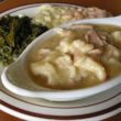 Oblong bowl of chicken and dumplings and gravy is backed by turnip greens, creamed corn, and beans.