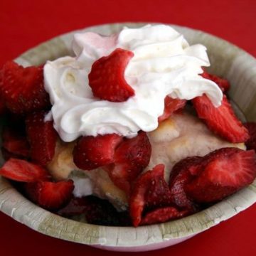 In a paper bowl, a baking powder biscuit is topped with strawberries and whipped cream.
