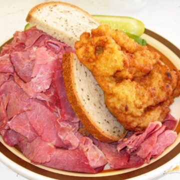 Brick-red corned beef spills out all sides of the rye bread that holds it. Atop this sandwich is a crusty potato pancake.