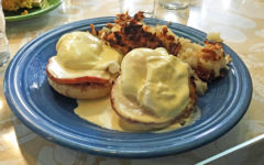 Traditional eggs Benedict, topped with hollandaise, perched upon Canadian bacon and English muffin halves. Hash browns in the background