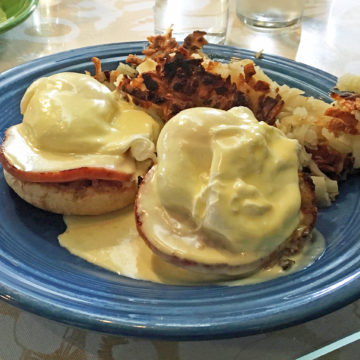 Traditional eggs Benedict, topped with hollandaise, perched upon Canadian bacon and English muffin halves. Hash browns in the background