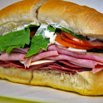 Cold cuts are piled high with garnishes and condiments in a long loaf almost too big to lift