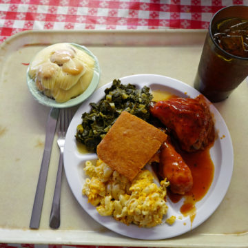 Cafeteria tray holds a square meal of BBQ chicken, mac & cheese, collards, cornbread, sweet tea, and banana pudding