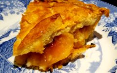 Peach slices peek out from under the rugged pastry crust of a pie slice on a china plate.