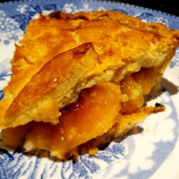 Peach slices peek out from under the rugged pastry crust of a pie slice on a china plate.