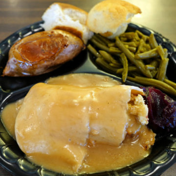 Disposable partitioned plate holds turkey with dressing under gravy, a yam, green beans, cranberry sauce, and a baked yam