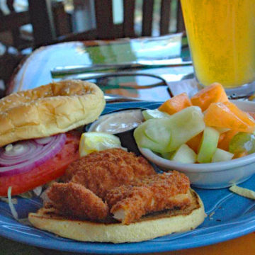 Great hunks of golden fried hogfish perch on the bottom of a bun, accompanied by garnishes and a bowl of fresh-cut fruit.