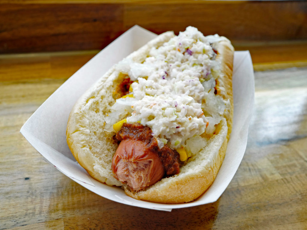 Cardboard boat holds a bunned hot dog dressed with chili, slaw, and mustard.
