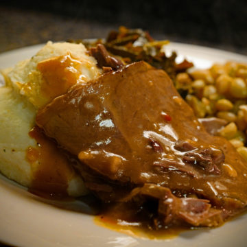 Thick-cut roast beef covered with gravy, on a plate with mashed potatoes and beans