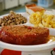 On a partitioned plate, a slab of sweet-sauced meatloaf accompanied by black-eyed peas, mac & cheese, and cornbread