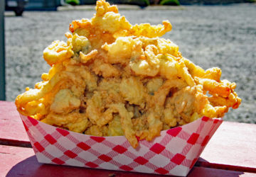 On a sunny picnic table, fried clams are piled high in a red-checked cardboard boat.