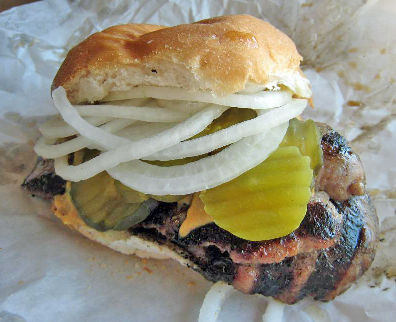 Charcoal-grilled sausage is piled into a bun with pickle chips and onions.