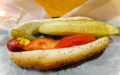 Poppy-seed bun holds hot dog with mustard, tomato, relish, pickle spear, and sport pepper