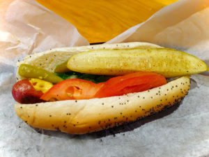 Poppy-seed bun holds hot dog with mustard, tomato, relish, pickle spear, and sport pepper