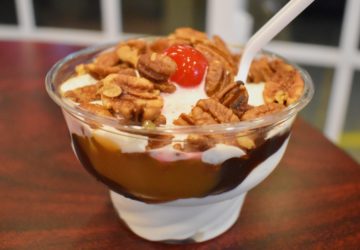 In a clear plastic cup, ice cream is topped with swirls of hot fudge and caramel, pecan halves, and a maraschino cherry
