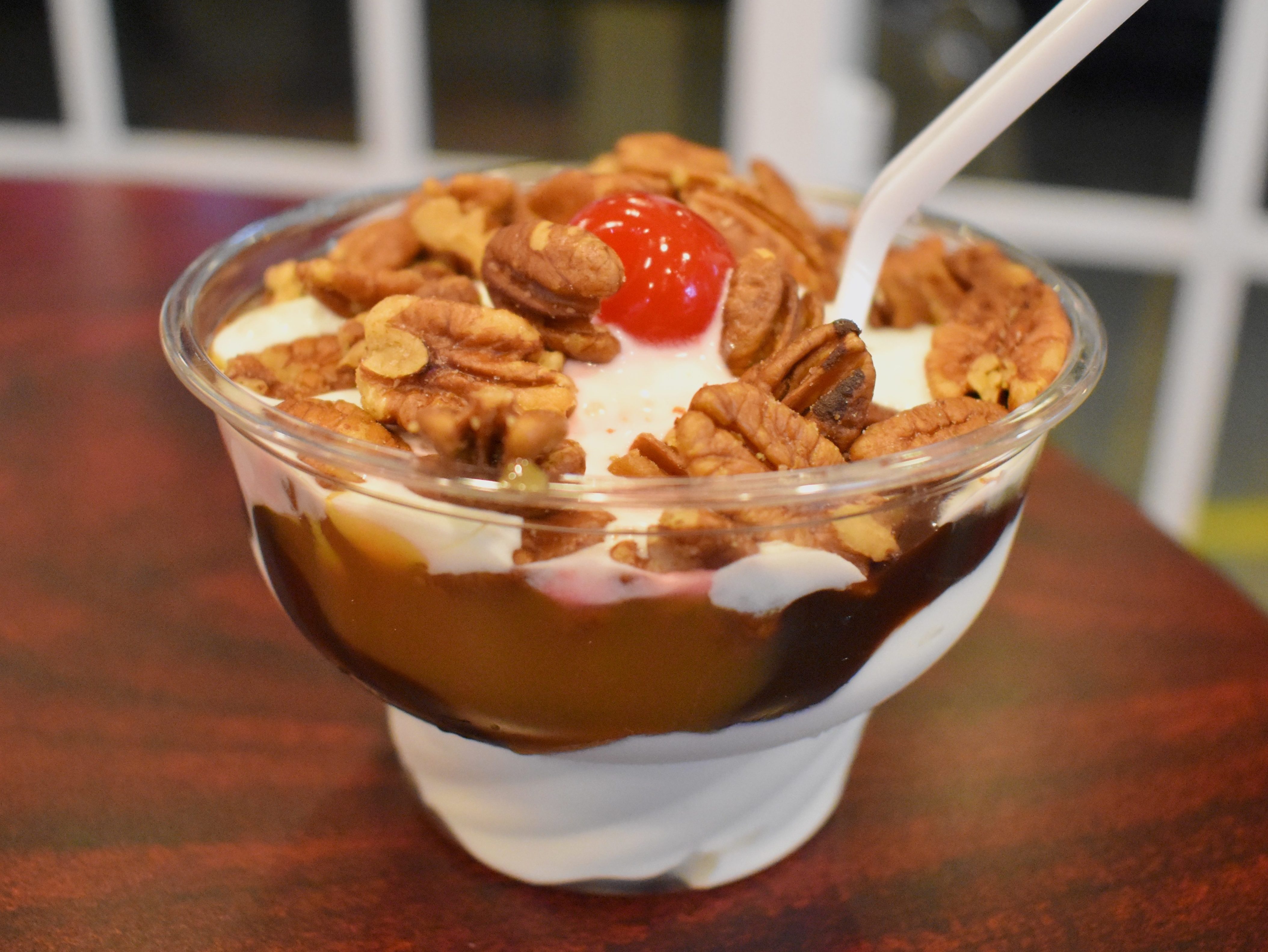 In a clear plastic cup, ice cream is topped with swirls of hot fudge and caramel, pecan halves, and a maraschino cherry