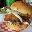 Big hunk of golden-fried chicken is sandwiches with cabbage slaw and pickles, with a cup of cucumber slaw in the background.