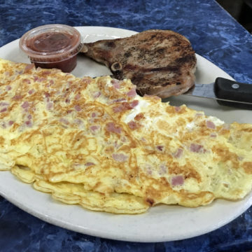 Huge ham & cheese omelet on a plate sided by a a pork chop, which looks small in comparison