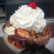 Glass bowl holds vanilla ice cream topped with chocolate & caramel sauces, chopped nuts, whipped cream, and a cherry.