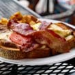 Plate on an outdoor table holds a hot brown sandwich criss-crossed with bacon