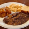 On a disposable partitioned plate, gravy-sopped pot roast atop a bed of mashed potatoes is accompanied by rutabagas, mac & cheese, and a block of cornbread