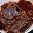 Bowl of thin pieces of burnished-red beef jerky show hash marks from air-drying on a grate.
