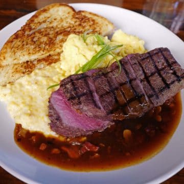 Breakfast plate includes sliced steak, scrambled eggs, and toast, atop a spill of mushroom gravy